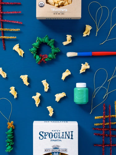 Pasta shaped like Christmas trees and wreath decorations on a blue background with Sfoglini pasta packaging.