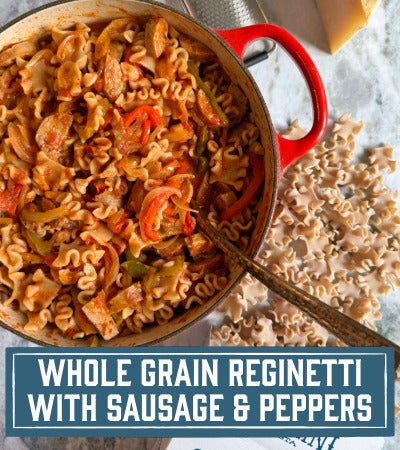 Red pot filled with pasta dish labeled 'Whole Grain Reginetti with Sausage & Peppers' on a light background.