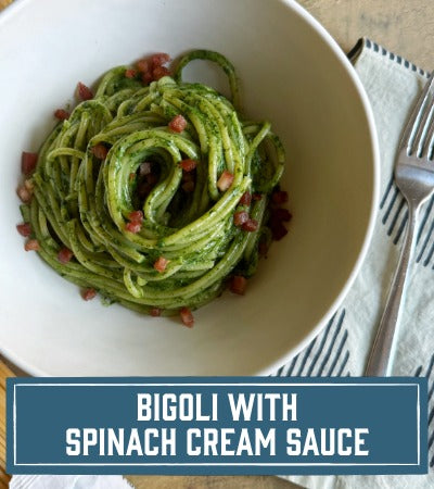 Sfoglini Pasta Recipe: Green long pasta noodles dish with spinach cream sauce in a white bowl on a wooden table. With text overlay "Bigoli with Spinach Cream Sauce"