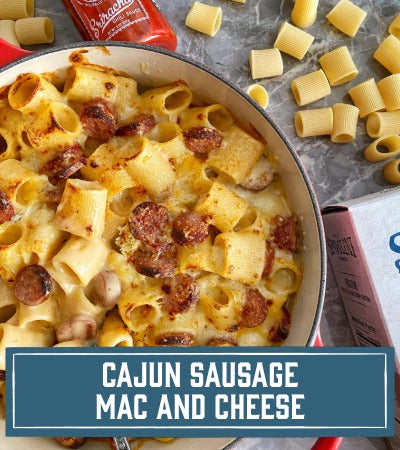 Sfoglini Pasta Recipe:  Cajun sausage mac and cheese in a pan with Rigatoni pasta and cheese on a table. with text overlay "Cajun Sausage Mac and Cheese"