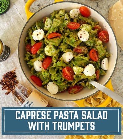 Sfoglini Pasta Recipe:  Caprese pasta salad with Trumpets, tomatoes, mozzarella, and pesto in a white bowl on a wooden surface. With text overlay "Caprese Pasta Salad with trumpets"