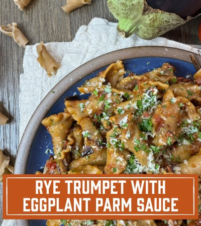 Sfoglini Pasta Recipe: Pasta dish with eggplant Parm sauce on a plate, garnished with herbs, on a wooden surface. With text overlay "Rye Trumpet with Eggplant Parm Sauce"