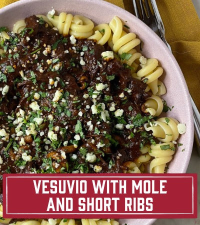 Sfoglini Pasta Recipe: Bowl of pasta with mole and short ribs, garnished with green herbs, on a yellow cloth background. With text overlay " Vesuvio with Mole and Short Ribs"