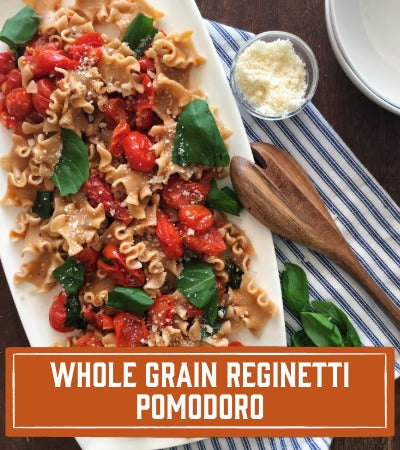 Sfoglini Pasta Recipe: Pasta dish with tomatoes and basil on a striped tablecloth, with a wooden spoon and small bowl of cheese. With text overlay " Whole Grain Reginetti Pomodoro"