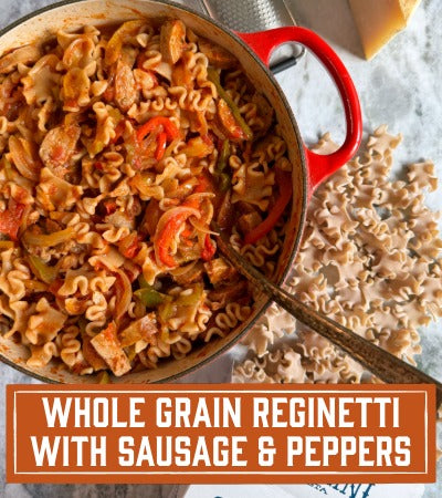 Sfoglini Pasta Recipe: Red pot filled with pasta dish on a marble surface. With text overlay "Whole Grain Reginetti with Sausage & Peppers"