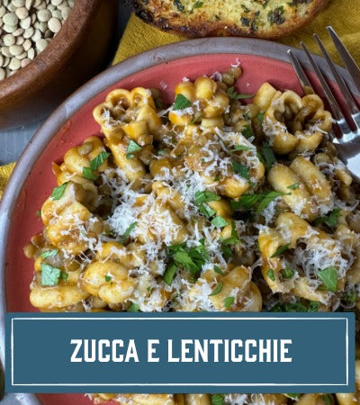 Sfoglini Pasta Recipe: Plate of Zucca pasta with lentils, garnished with herbs and cheese, on a red plate with a fork. With text overlay "Zucca E Lenticchie"