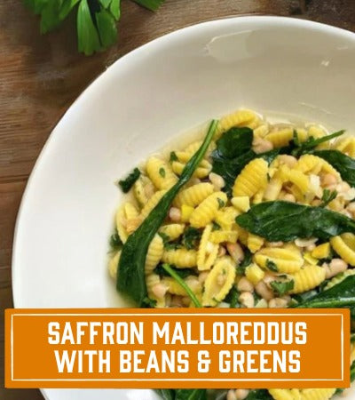 Sfoglini Pasta Recipe: White bowl of saffron malloreddous with beans, broth, and greens on a wooden surface. With text overlay "saffron malloreddous with beans and greens"