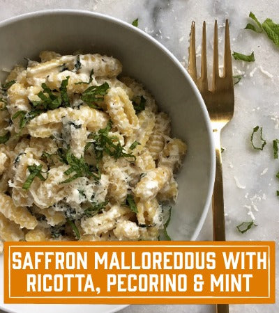Sfoglini Pasta Recipe: Bowl of saffron malloreddous with ricotta, pecorino, and mint on a marble surface with a gold fork. with text overlay "saffron malloreddous with ricotta, pecorino, and mint"