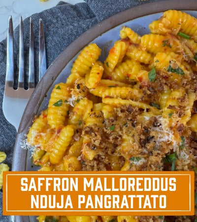 Sfoglini Pasta Recipe: Plate of saffron malloreddus with nduja pangrattato on a gray napkin with a fork. With text overlay "saffron malloreddus with nduja pangrattato"