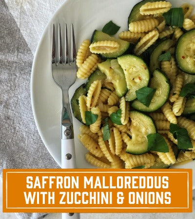 Sfoglini Pasta Recipe: Plate of saffron malloreddus with zucchini and onions on a white plate with a fork, on a light fabric background. With textt overlay "saffron malloreddus with zucchini and onions"