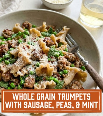 Pasta dish with sausage, peas, and mint in a white bowl on a light surface. With text "Whole Grain Trumpets with Sausage, Peas, & Mint"