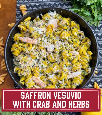 Sfoglini Pasta Recipe: Saffron Vesuvio with crab and Herbs. Black bowl filled with yellow pasta salad with chicken and herbs on a dark surface. With text overlay "Saffron Vesuvio with rab and Herbs"