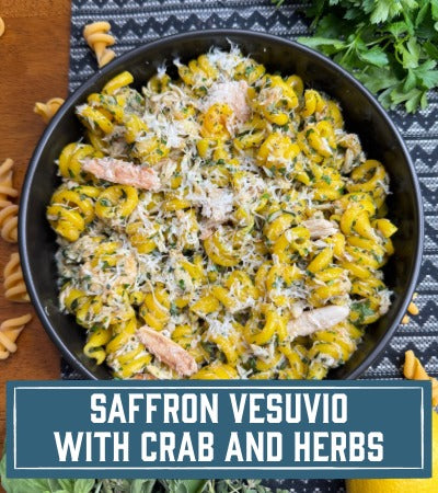 Sfoglini Pasta Recipe: Saffron Vesuvio with crab and Herbs. Black bowl filled with yellow pasta salad with chicken and herbs on a dark surface. With text overlay "Saffron Vesuvio with rab and Herbs"