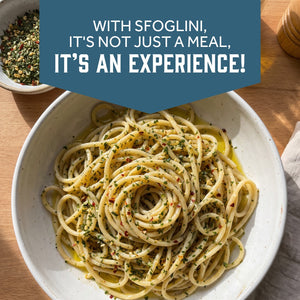 A top-down view of a white bowl filled with sfoglini bigoli pasta topped with herbs, oil, and red pepper flakes on a wooden table. With text  - WITH SFOGLINI, IT'S NOT JUST A MEAL, IT'S AN EXPERIENCE!