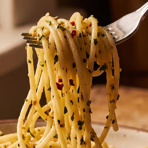 Close-up of sfoglini bigoli pasta tossed with herbs and red pepper flakes, lifted on a fork against a warm indoor background.